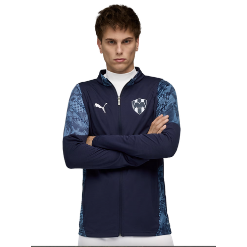 A young man with arms crossed wearing a navy Puma jacket with light blue patterned sleeves and a team crest, set against a black background.