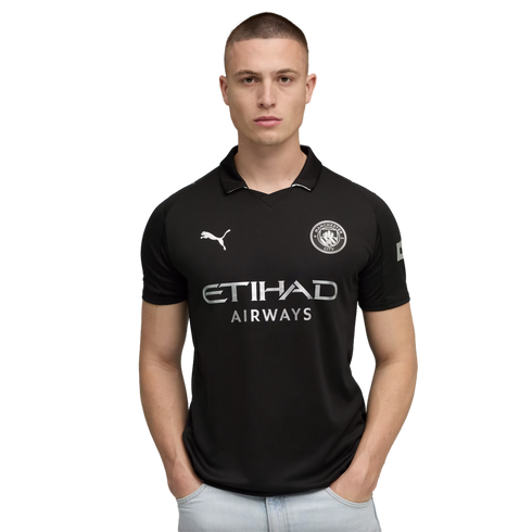 A man in a black Manchester City soccer jersey and light-wash jeans stands with his hands in his pockets against a solid black background.