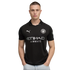 A man in a black Manchester City soccer jersey and light-wash jeans stands with his hands in his pockets against a solid black background.