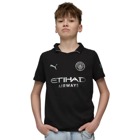 A young boy with light brown hair wears a black Manchester City soccer jersey and light blue jeans, posing with his hands in his pockets against a solid black background.
