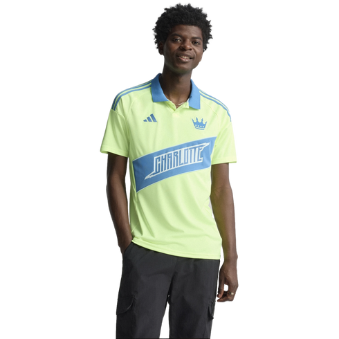 A smiling man wearing a neon yellow Charlotte FC polo shirt and black cargo pants against a black background.