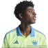 Profile of a smiling young Black man with an afro wearing a lime green Adidas polo shirt with a blue collar and crown logo, against a black background.