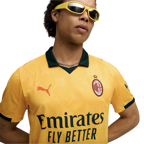 A man wearing a yellow AC Milan soccer jersey with a dark green collar and yellow-framed sunglasses against a solid black background.