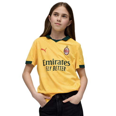A young girl with long brown hair poses in a yellow AC Milan football jersey and black pants against a black background.