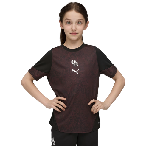 A young girl stands with hands on her hips wearing a maroon and black Puma athletic shirt against a solid black background.
