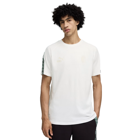 A curly-haired man stands against a black background wearing a white Puma short-sleeved t-shirt and dark trousers with patterned side details.