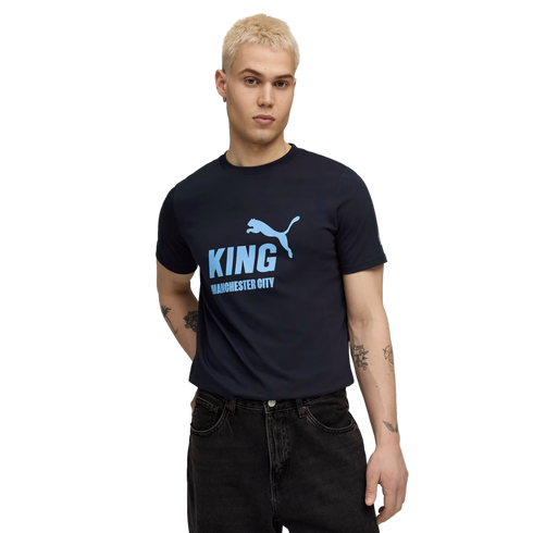A person with short blonde hair wears a dark blue Manchester City T-shirt and black jeans against a black background.