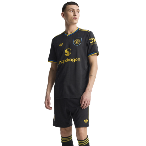 A man models the black Manchester United 2024/25 third kit, featuring yellow and blue trim, the Adidas Trefoil logo, and Snapdragon sponsor.