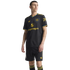 A man models the black Manchester United 2024/25 third kit, featuring yellow and blue trim, the Adidas Trefoil logo, and Snapdragon sponsor.
