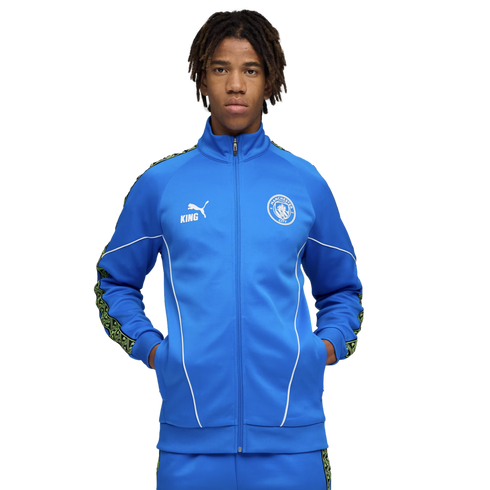 Young man with dreadlocks wearing a blue Manchester City tracksuit featuring a Puma logo and club crest, standing against a black background.