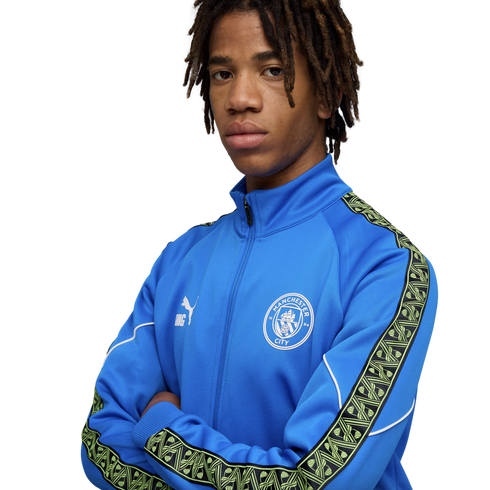 Young man with dreadlocks wearing a blue Manchester City tracksuit jacket, posing with arms crossed against a black background.