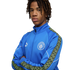 Young man with dreadlocks wearing a blue Manchester City tracksuit jacket, posing with arms crossed against a black background.