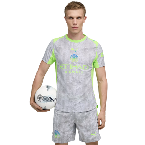 Man in a Manchester City soccer kit, grey with neon green accents, holding a soccer ball under his arm against a black background.