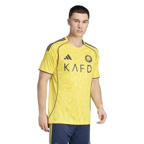 A man modeling a yellow soccer jersey with navy blue trim and "KAFD" across the chest, paired with navy blue shorts. The jersey features a subtle wavy pattern.