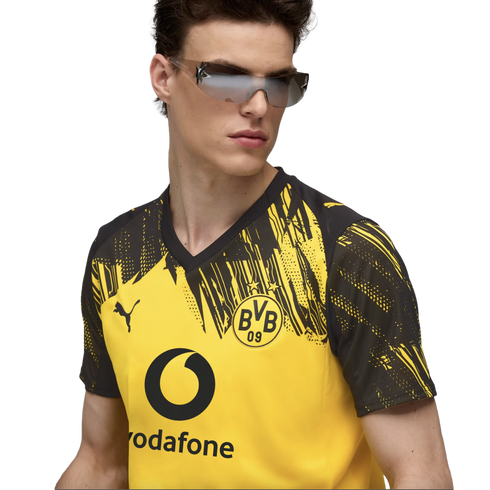 A man wears a yellow and black Puma BVB jersey and futuristic sunglasses against a black background.