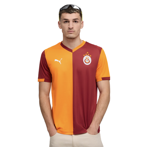 Man in an orange and maroon Galatasaray football jersey with a white Puma logo and team crest, standing against a black background with hands in pockets.