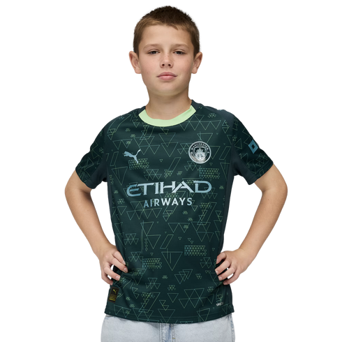 A young boy posing in a dark green Manchester City soccer jersey with geometric triangle patterns, hands on his hips against a black background.