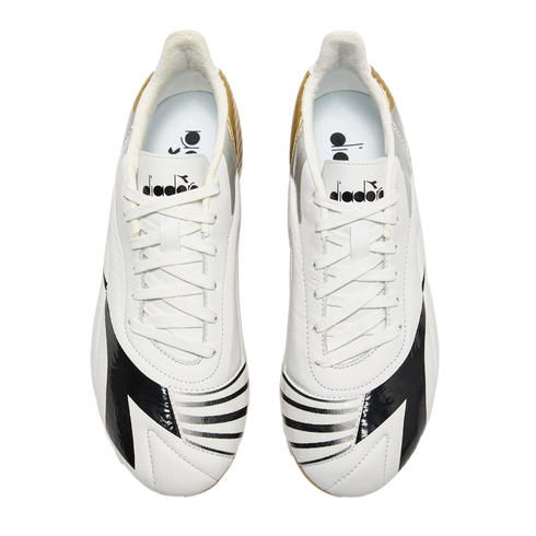 Top-down view of white Diadora soccer cleats with black graphic patterns on the toes and gold and silver accents on the heels, against a black background.