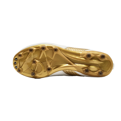 Bottom view of a white and gold soccer cleat featuring a metallic gold sole plate and circular studs, set against a solid black background.