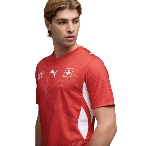 A man in a red Puma Switzerland national soccer jersey, looking to the side against a black background.