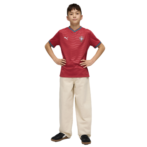 A boy posing in a red wavy-patterned sports jersey and cream-colored wide-leg trousers with his hands on his hips against a black background.