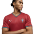 Man with braided hair in a red Portugal Puma soccer jersey, arms crossed, looking sideways against a black background.