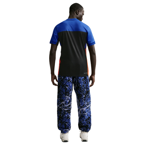 Back view of a man in a blue and black t-shirt and blue marbled print trousers with white sneakers, against a solid black background.