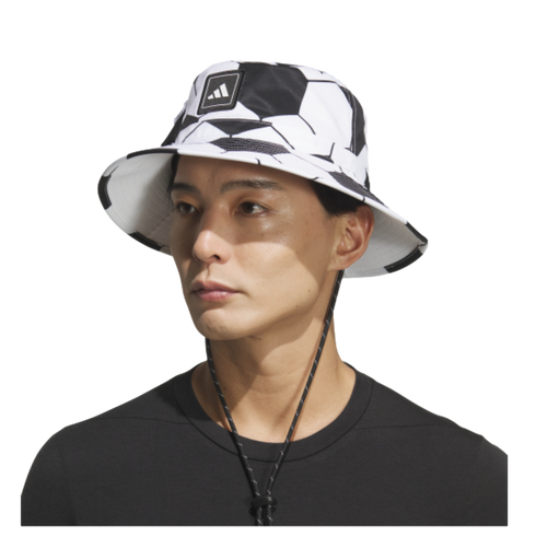 A man wears a white Adidas bucket hat with a black soccer ball pattern and a chin strap, set against a black background.