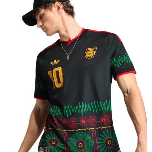 Man in a black Jamaica football jersey with vibrant green and red graphic patterns, a gold number 10, and team crest. He wears a black cap and silver necklace.