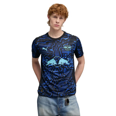 A man with red hair wearing a black and blue patterned Puma RB Leipzig soccer jersey and light blue jeans.