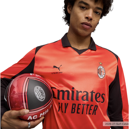 A man wearing an orange Puma AC Milan soccer jersey holds a matching soccer ball against a black background.