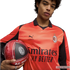 A man wearing an orange Puma AC Milan soccer jersey holds a matching soccer ball against a black background.
