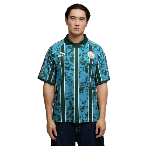 Man wearing a teal and dark green patterned Manchester City soccer jersey with vertical stripes, posed against a plain black background.