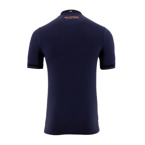 A back view of a navy blue short-sleeved soccer jersey against a solid black background. Near the collar, there is a small Colombian flag above the copper-colored Spanish text, "SOÑAR TRASCENDER Y HACER HISTORIA." The sleeve cuffs are accented with a matc