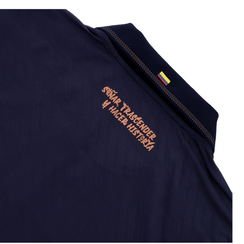 A close-up of the back collar area of a dark navy blue shirt. Below the collar, a Spanish slogan is printed in a stylized copper-colored font that reads "SOÑAR, TRASCENDER Y HACER HISTORIA." A small Colombian flag is visible on the ribbed collar, and the