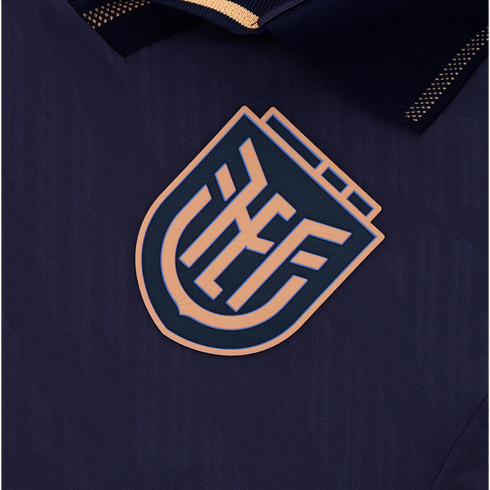 A close-up of a dark navy blue athletic jersey featuring a raised, shield-shaped logo with stylized, interlocking tan letters. The jersey fabric has a subtle wavy texture, and a dark collar with gold accents is partially visible at the top.