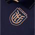 A close-up of a dark navy blue athletic jersey featuring a raised, shield-shaped logo with stylized, interlocking tan letters. The jersey fabric has a subtle wavy texture, and a dark collar with gold accents is partially visible at the top.