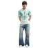 A young man with dark, textured hair stands against a solid black background. He is wearing a light green and teal patterned athletic jersey with a crest and the number 7, wide-leg light-wash blue jeans, and white sneakers. He stands with his hands behind