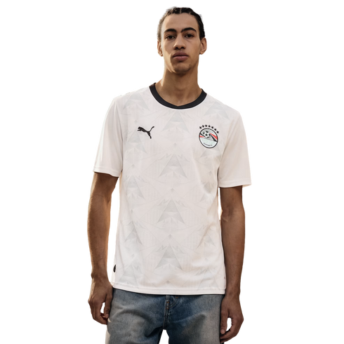A young man wearing a white Puma Egypt national football team away jersey with a black collar and a tonal geometric pattern, paired with blue jeans against a solid black background.