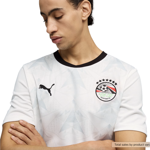 A young man looks down with his arms crossed, wearing a white Puma soccer jersey with a black crew neck and a subtle geometric pattern. The jersey features the Egyptian Football Association crest on the left chest and a black Puma logo on the right, all s