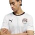 A young man looks down with his arms crossed, wearing a white Puma soccer jersey with a black crew neck and a subtle geometric pattern. The jersey features the Egyptian Football Association crest on the left chest and a black Puma logo on the right, all s