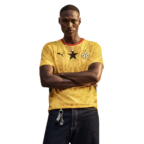 A man with his arms crossed stands against a black background, wearing a yellow Ghana national team soccer jersey and dark jeans.
