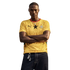 A man with his arms crossed stands against a black background, wearing a yellow Ghana national team soccer jersey and dark jeans.