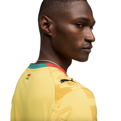 A profile view of a Black man looking over his shoulder, wearing a yellow Puma sports jersey with the flag of Ghana on the back of the neck and a patterned design on the shoulder, set against a solid black background.