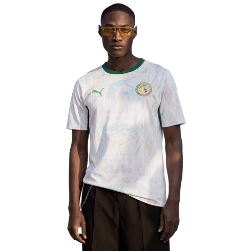 A man wearing yellow-tinted sunglasses and a white Senegal national team football jersey with a green collar, standing against a solid black background.