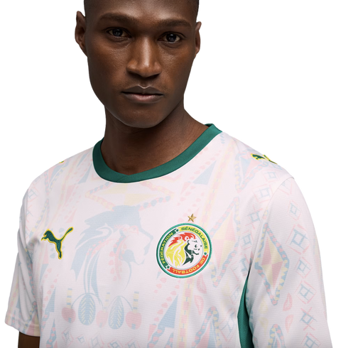 A man in a white Senegal national team soccer jersey with green trim and faint colorful geometric patterns, set against a solid black background.