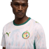 A man in a white Senegal national team soccer jersey with green trim and faint colorful geometric patterns, set against a solid black background.
