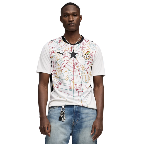 A medium shot of a Black man wearing a white Puma Ghana national football team jersey with a colorful geometric pattern and a central black star. He is also wearing light-wash blue jeans and a silver necklace, set against a solid black background.