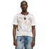 A medium shot of a Black man wearing a white Puma Ghana national football team jersey with a colorful geometric pattern and a central black star. He is also wearing light-wash blue jeans and a silver necklace, set against a solid black background.