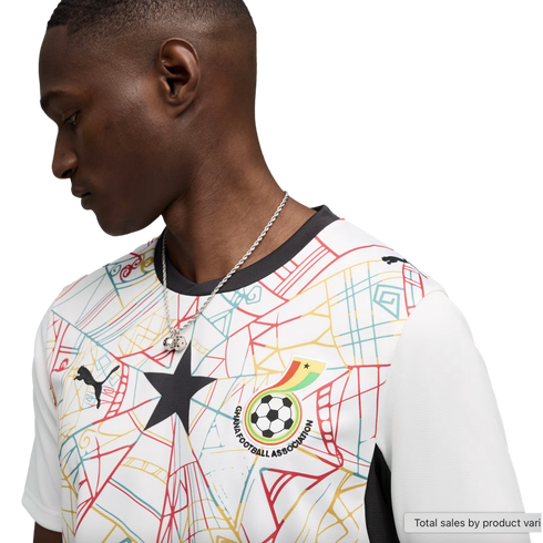 A side profile of a Black man wearing a white Ghana national team soccer jersey. The jersey features a colorful geometric pattern, a large black star in the center, and the Ghana Football Association crest. He is wearing a silver chain and looking downwar